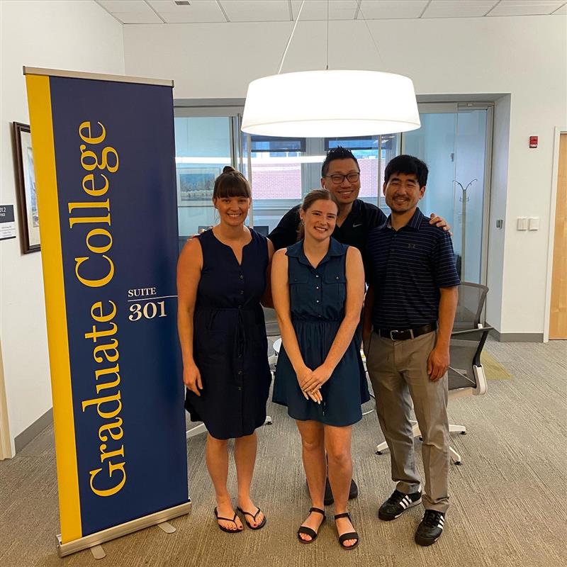 Image of Rachel Mroz, featured in the center with other staff members in the Graduate College. Also in the image is the Grad College vertical sign.